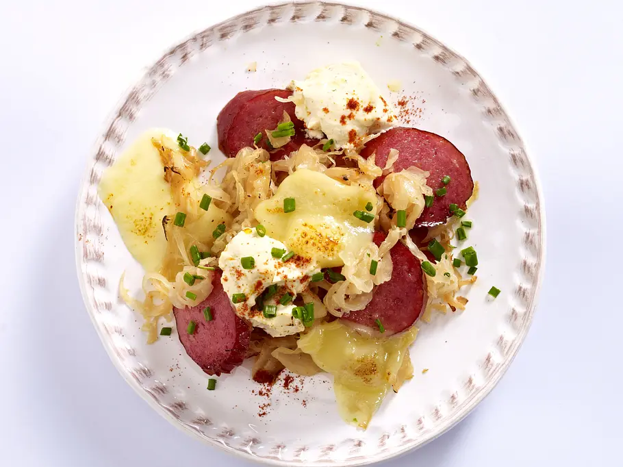 Raclette-Idee Weihnachten: Auf einem großen weißen Teller liegen in Scheiben geschnittene Cabanossi und Sauerkraut, überbacken mit Raclettekäse, garniert mit einem Klecks Paprika-Frischkäse und frischem Schnittlauch