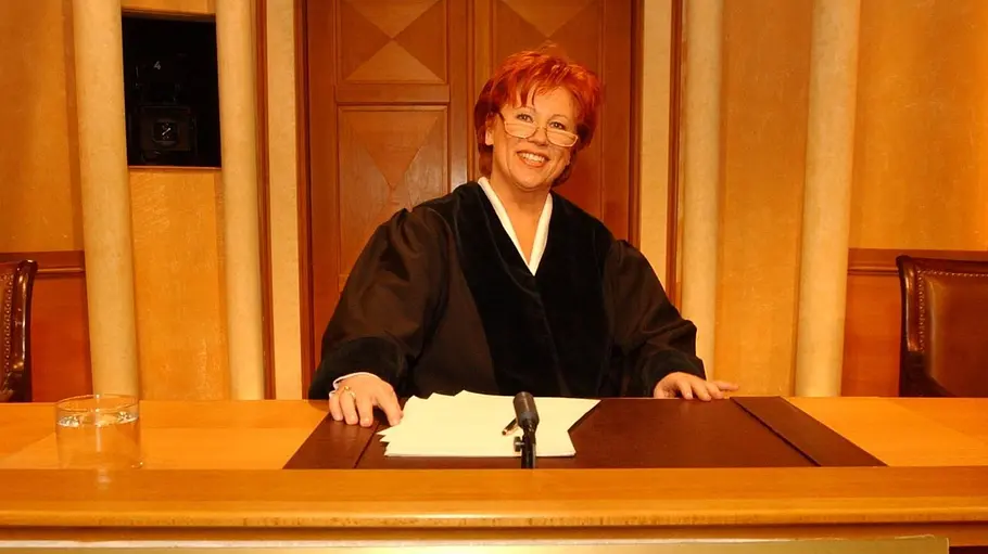 Barbara Salesch sitzt im Gerichtssaal am Richterpult und lächelt in die Kamera. Sie trägt eine Brille, vor ihr liegen mehrere Blätter Papier und ein goldenes Namensschild Richterin Barbara Salesch.