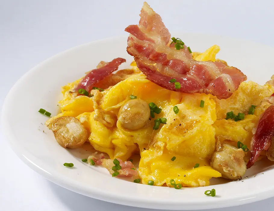 Rührei mit Speck und Champignons Ein weißer Teller mit Rührei, Champignons und gebratenen Speckscheiben. Mit Schnittlauch garniert.