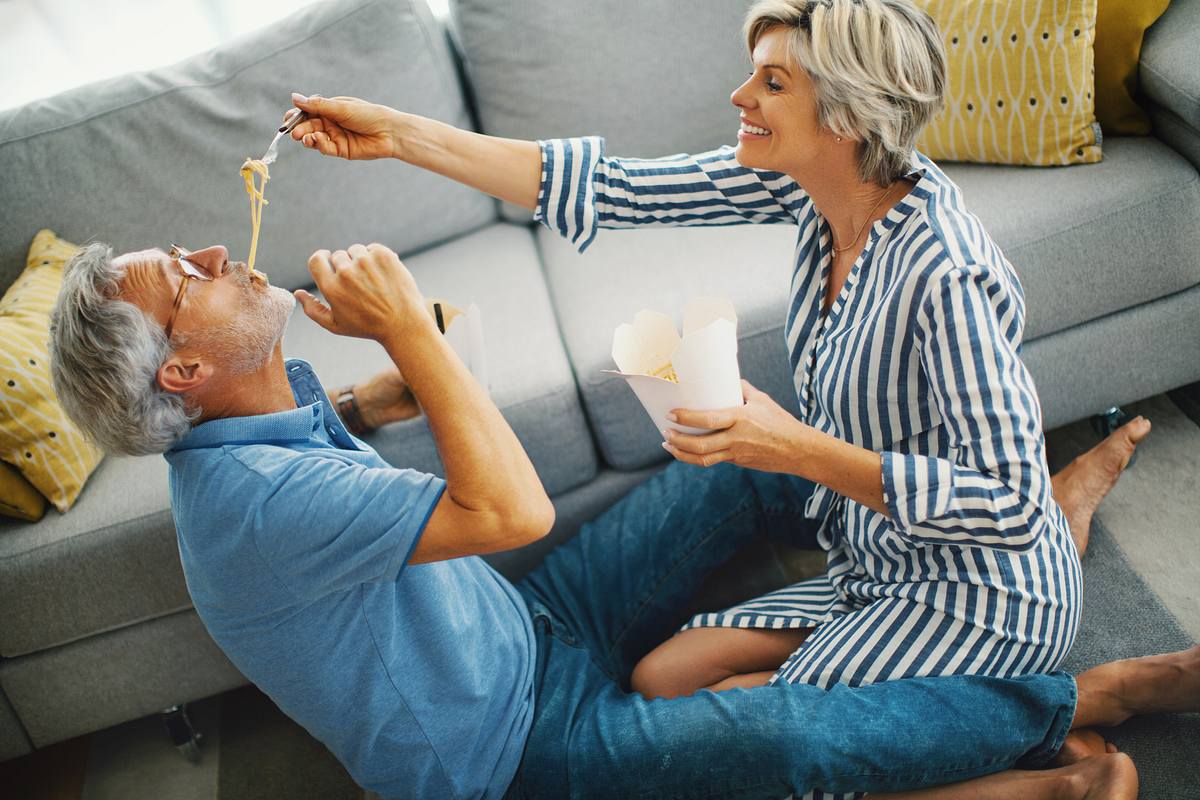 Mann und Frau teilen einen vertrauten Moment. Sie sitzen auf dem Boden, lachen und die Frau füttert den Mann mit asiatischem essen.