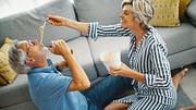 Mann und Frau teilen einen vertrauten Moment. Sie sitzen auf dem Boden, lachen und die Frau füttert den Mann mit asiatischem essen. - Foto: Aja Koska / iStock