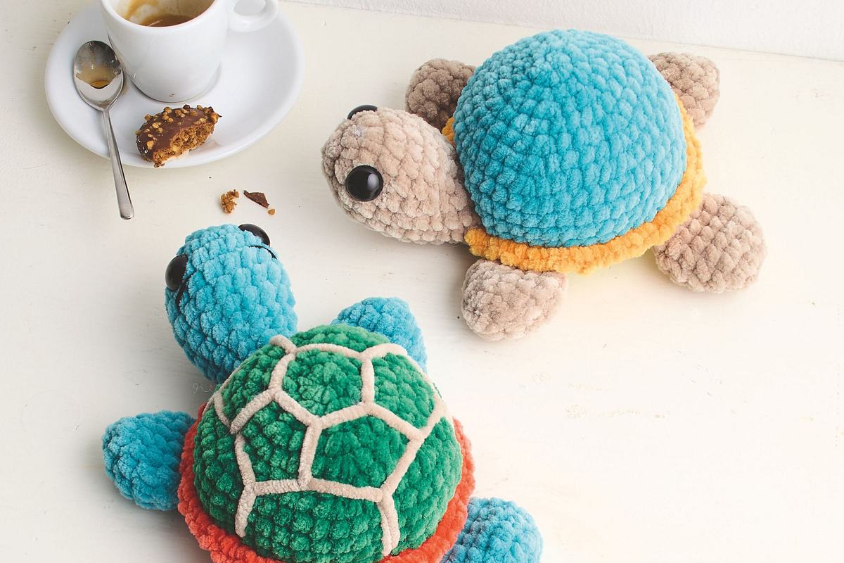 Zwei gehäkelte, flauschige Schildkröten, die auf eine kleine Espressotasse links oben im Bild zulaufen, um sich einen angeknabberten Keks zu schnappen, der auf dem Espressotellerchen liegt.