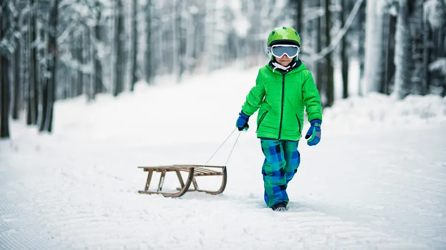 Schlitten für Kinder Kleiner Junge mit Helm und neongrünem Schneeanzug zieht einen Holzschlitten durch den Wald.