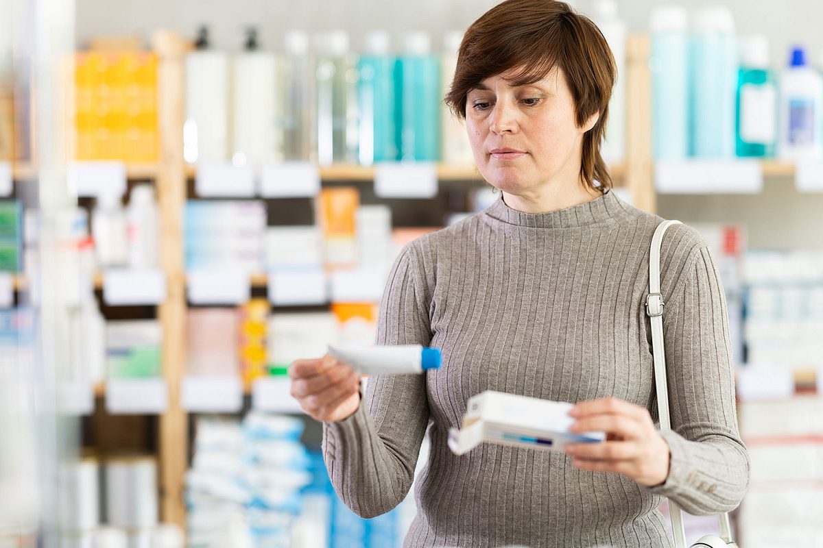 Frau steht in der Apotheke und betrachtet eine Salbe in ihrer Hand. (Themenbild)