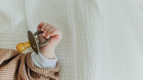 Kind hält einen Schnuller in der Hand auf einer weißen Decke. - Foto: Irina Brester/iStock