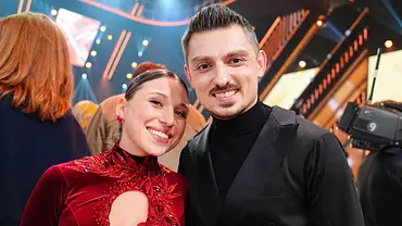 Ein Tanzpaar posiert lächelnd hinter den Kulissen von „Let’s Dance“; sie trägt ein rotes Bühnenkleid mit Glitzer, er ein schwarzes Outfit, im Hintergrund ist das TV-Studio zu sehen. - Foto: RTL / Stefan Gregorowius Ein Tanzpaar posiert lächelnd hinter den Kulissen von „Let’s Dance“; sie trägt ein rotes Bühnenkleid mit Glitzer, er ein schwarzes Outfit, im Hintergrund ist das TV-Studio zu sehen. - Foto: RTL / Stefan Gregorowius