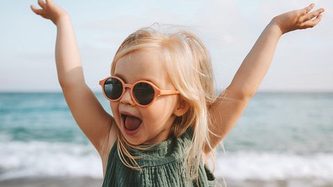 Mädchen mit Sonnenbrille wirft am Strand die Hände in die Luft (Themenbild) - Foto: Everste/iStock