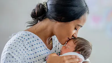 Eine Mutter hält ihr Neugeborenes im Arm und küsst es auf die Stirn. - Foto: iStock/FatCamera