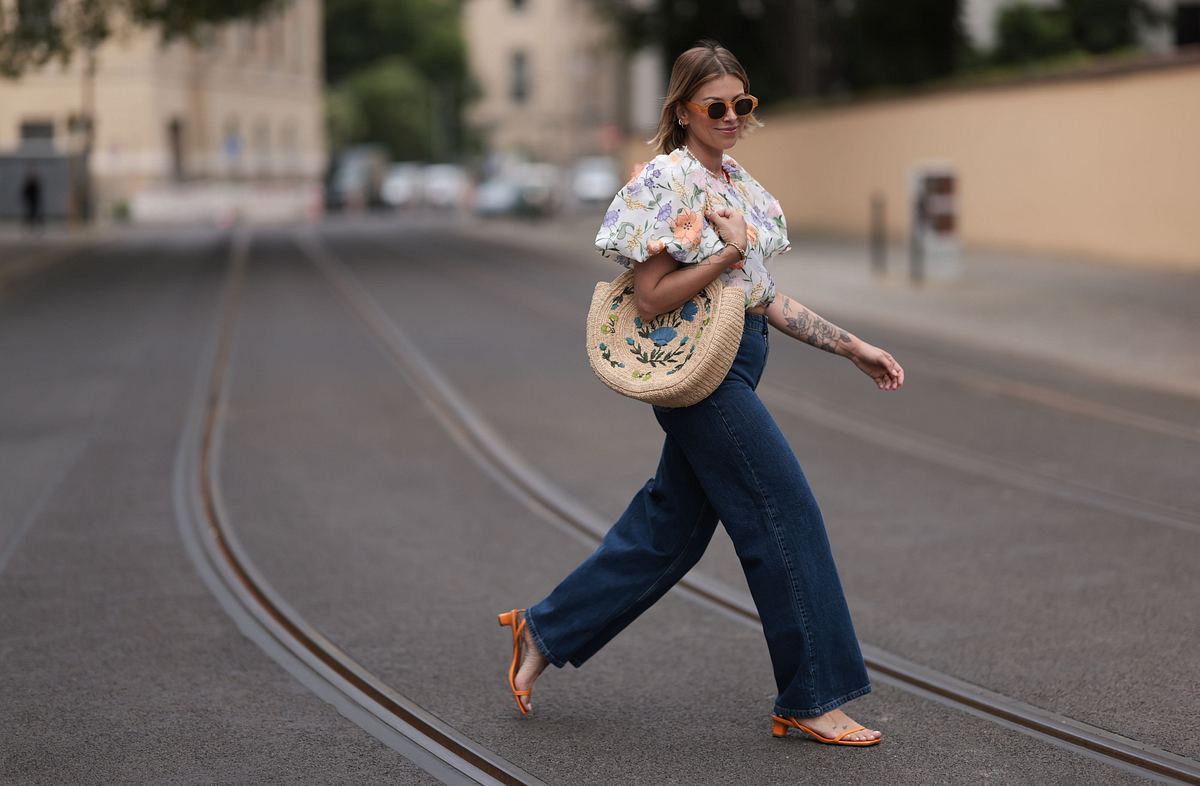 Eine Frau trägt eine weite Jeans, eine Strohtasche und eine luftige Bluse und läuft über die Straße. 