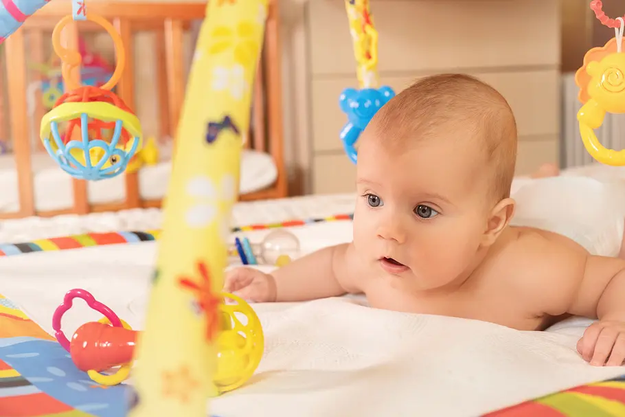 Spielbogen fördert die Entwicklung deines Babys – eine Expertin klärt auf Baby liegt bäuchlings unter einem Spielbogen und entdeckt die Spielgeräte