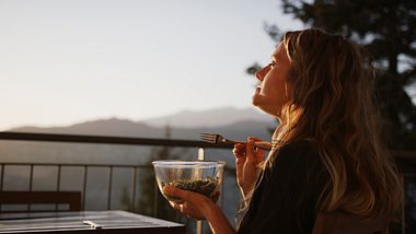 Frau sitzt mit Essen auf dem Balkon und genießt die Ruhe - Foto: AscentXmedia/iStock