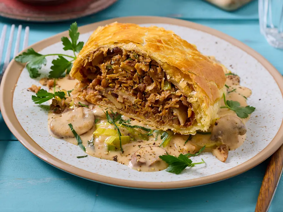 Eine Scheibe Blätterteigstrudel mit brauner Linsenfüllung auf Pilzsoße mit Petersilie.