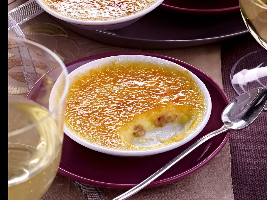 Goldbraune Crème-Brûlée in einer weißen flachen Schüssel mit einem Glas Weißwein im Vordergrund.