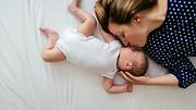 Frau liegt mit einem Baby auf dem Bett und küsst es auf die Nase (Themenbild) - Foto: AleksandarNakic/iStock