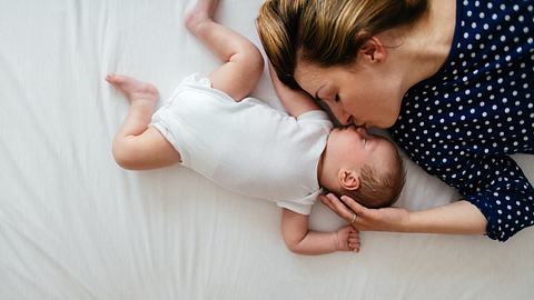 Frau liegt mit einem Baby auf dem Bett und küsst es auf die Nase (Themenbild) - Foto: AleksandarNakic/iStock