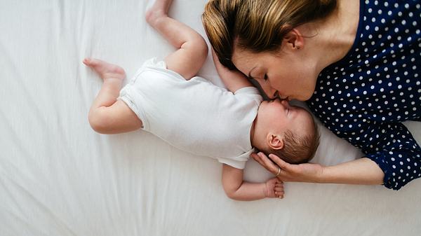 Frau liegt mit einem Baby auf dem Bett und küsst es auf die Nase (Themenbild) - Foto: AleksandarNakic/iStock
