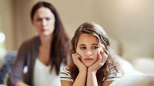 Ein Kind stützt die Hände in den Kopf und schaut traurig. Eine Mutter sitzt dahinter. - Foto: laflor / iStock