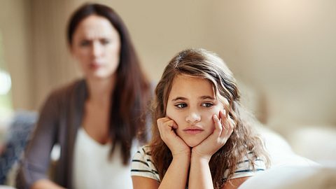 Ein Kind stützt die Hände in den Kopf und schaut traurig. Eine Mutter sitzt dahinter. - Foto: laflor / iStock