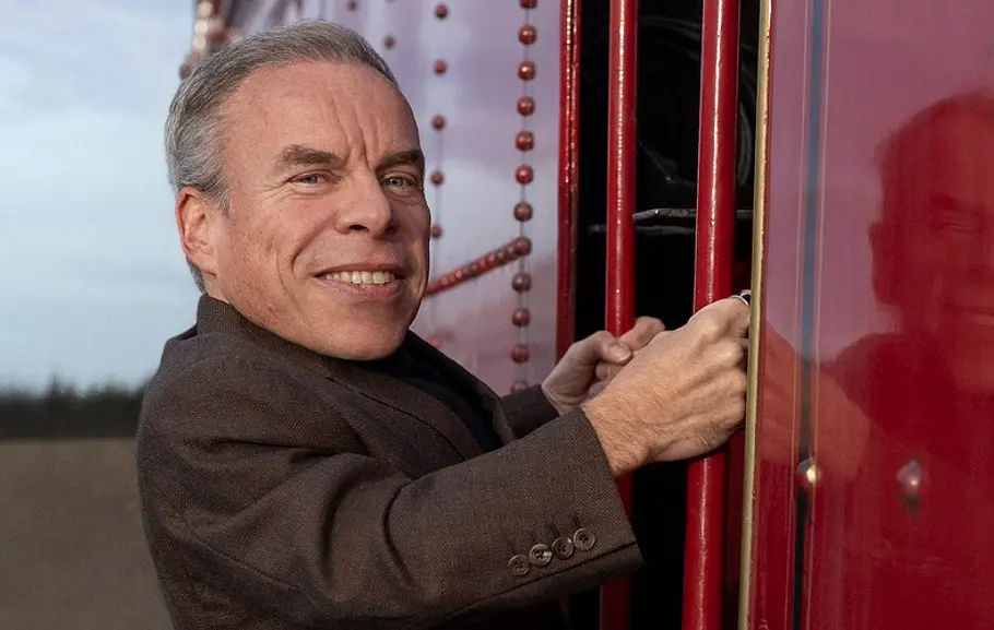 Warwick Davis Der Schauspieler Warwick Davis, ein Mann mit grauen Haaren und braunem Sakko hält sich an der Außenseite eines roten Zugwagens fest und lächelt in die Kamera. Seine Spiegelung ist auf der glänzenden Zugwand zu sehen. Im Hintergrund ist ein bewölkter Himmel und unscharfe Landschaft zu erkennen.