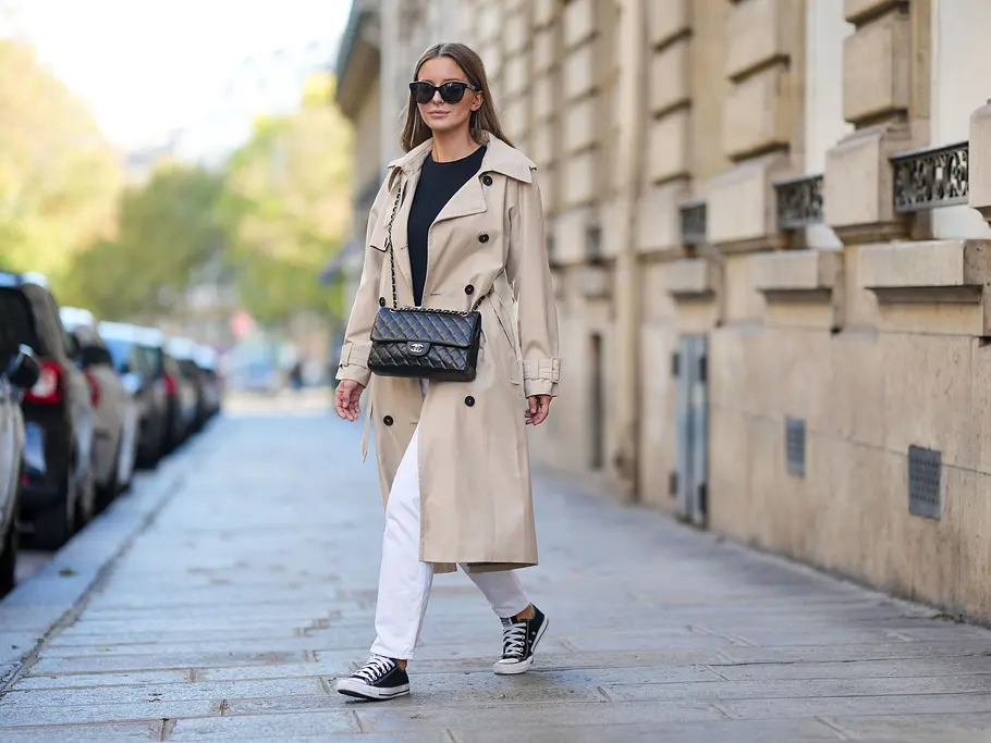 Weiße Jeans mit Trenchcoat kombinieren Weiße Straight Leg-Jeans mit einem schwarzen Oberteil, darüber ein klassischer beiger Trenchcoat und eine elegante schwarze Handtasche. Schwarze Halbschuh-Chucks runden das Outfit ab.