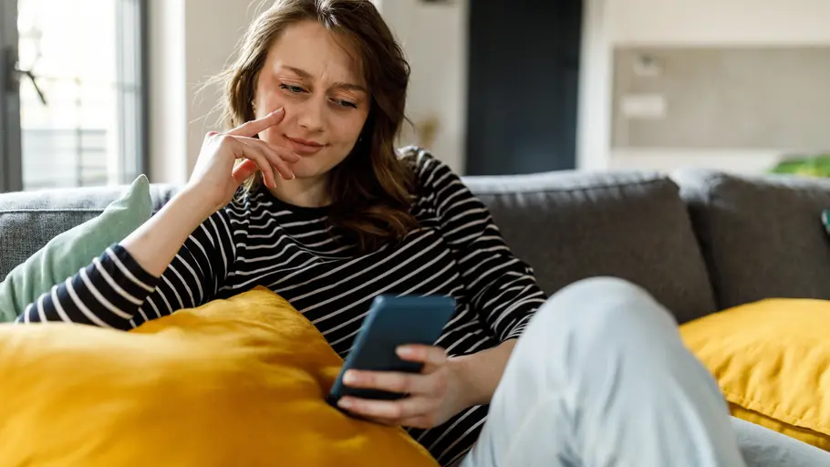 Neue WhatsApp-Betrugsmasche Frau sitzt auf dem Sofa und schaut skeptisch auf ihr Handy.