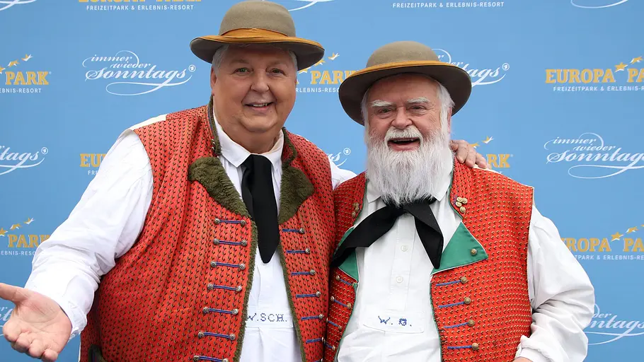 Wolfgang und Wilfried: Die originalen Wildecker Herzbuben Wolfgang Schwalm und Wilfried Gliem posieren gemeinsam in traditionellen roten Westen und Hüten vor einer Eventwand