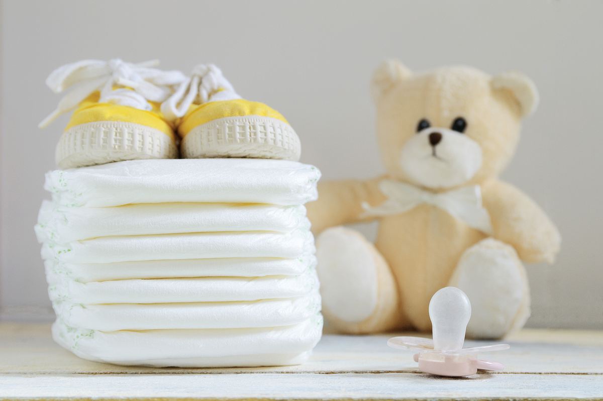 Ein Stapel Windeln neben einem Plüschteddy, gelben Babyschuhen und einem Babyschnuller