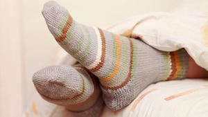 Bunt geringelte Wollsocken schauen unter einer Bettdecke hervor (Themenbild) - Foto: Alex_Doubovitsky/iStock