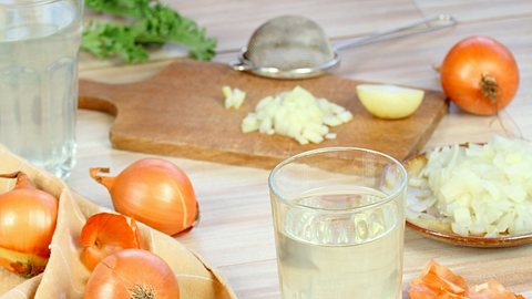 Ein Glas mit Zwiebeltee, Zwiebeln, Zwiebelschalen und geschnittenen Zwiebeln steht auf einem Tisch (Themenbild) - Foto: svehlik/iStock